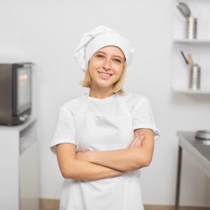 Horizontal shot of smiling attractive young woman, confectionery owner, posing to camera in her white uniform, hat and apron, standing in modern kitchen